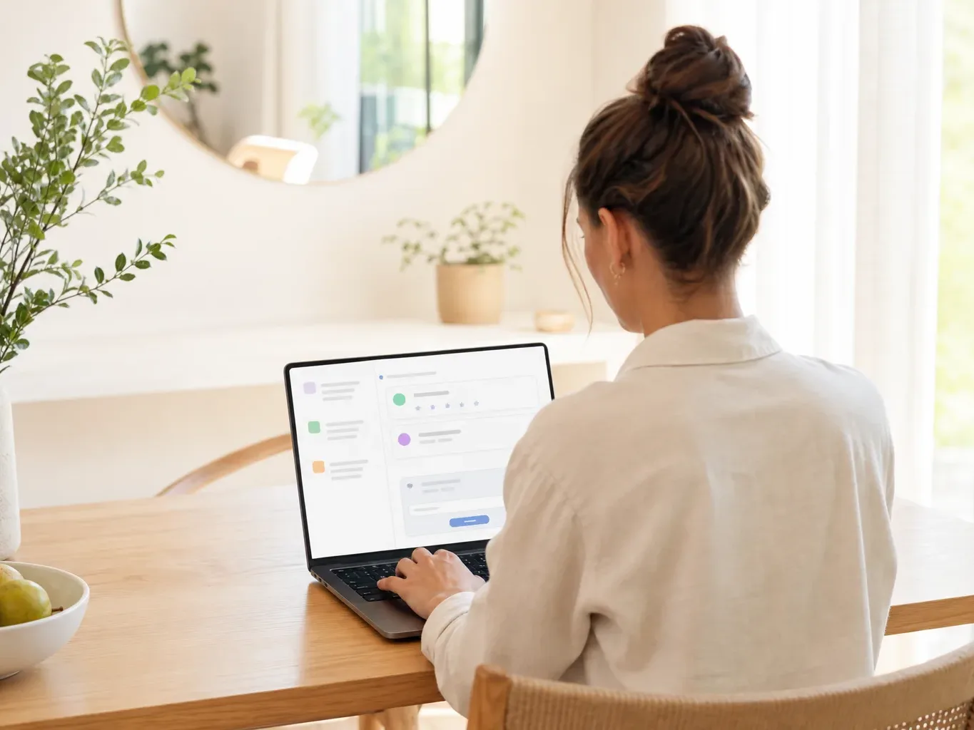 A person seen from behind sits at a wooden dining table in a bright home interior, typing on a laptop displaying a colourful abstract interface