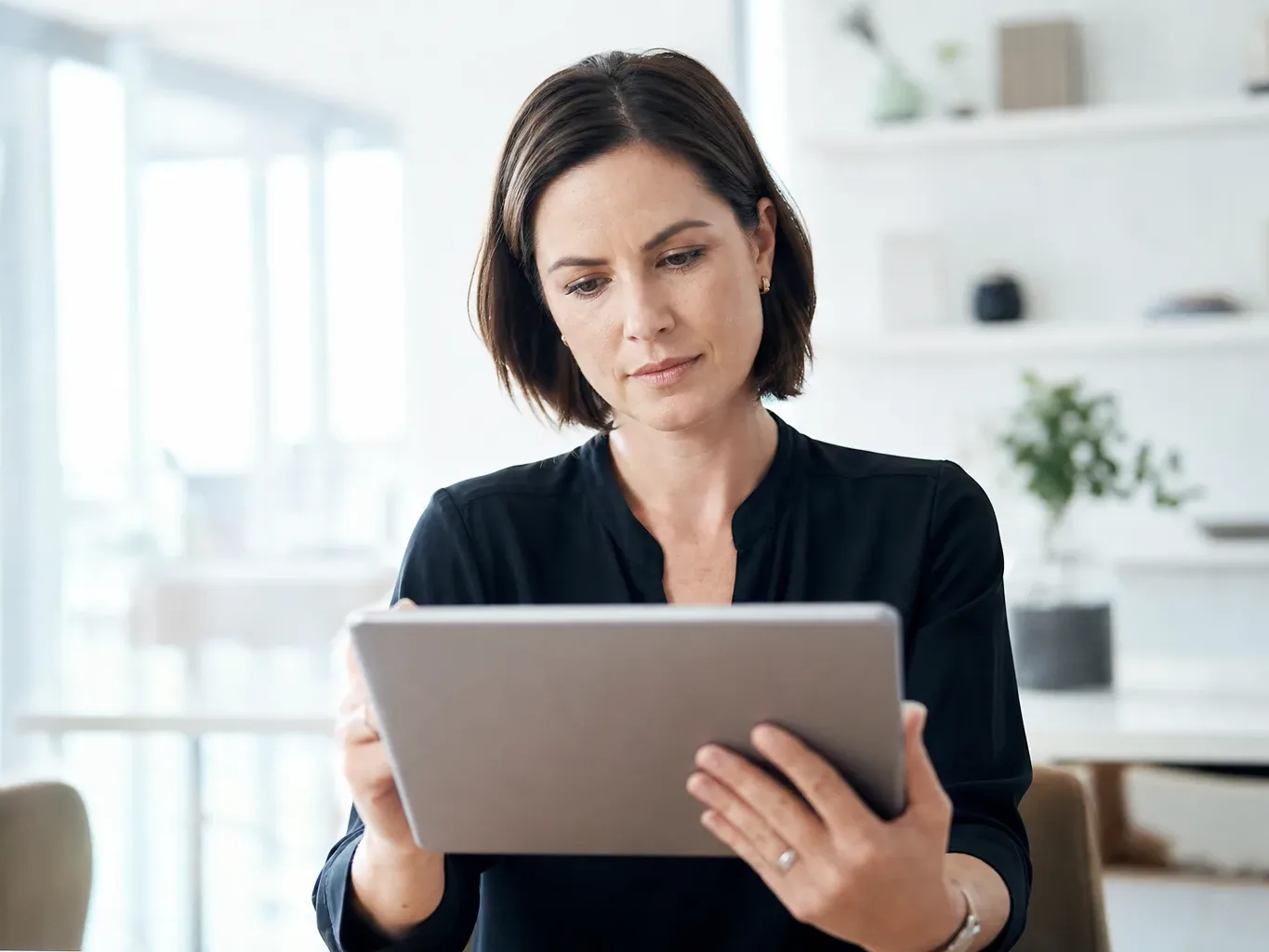 A person in a dark top stands indoors holding a tablet and reading its screen, with a softly blurred interior behind them