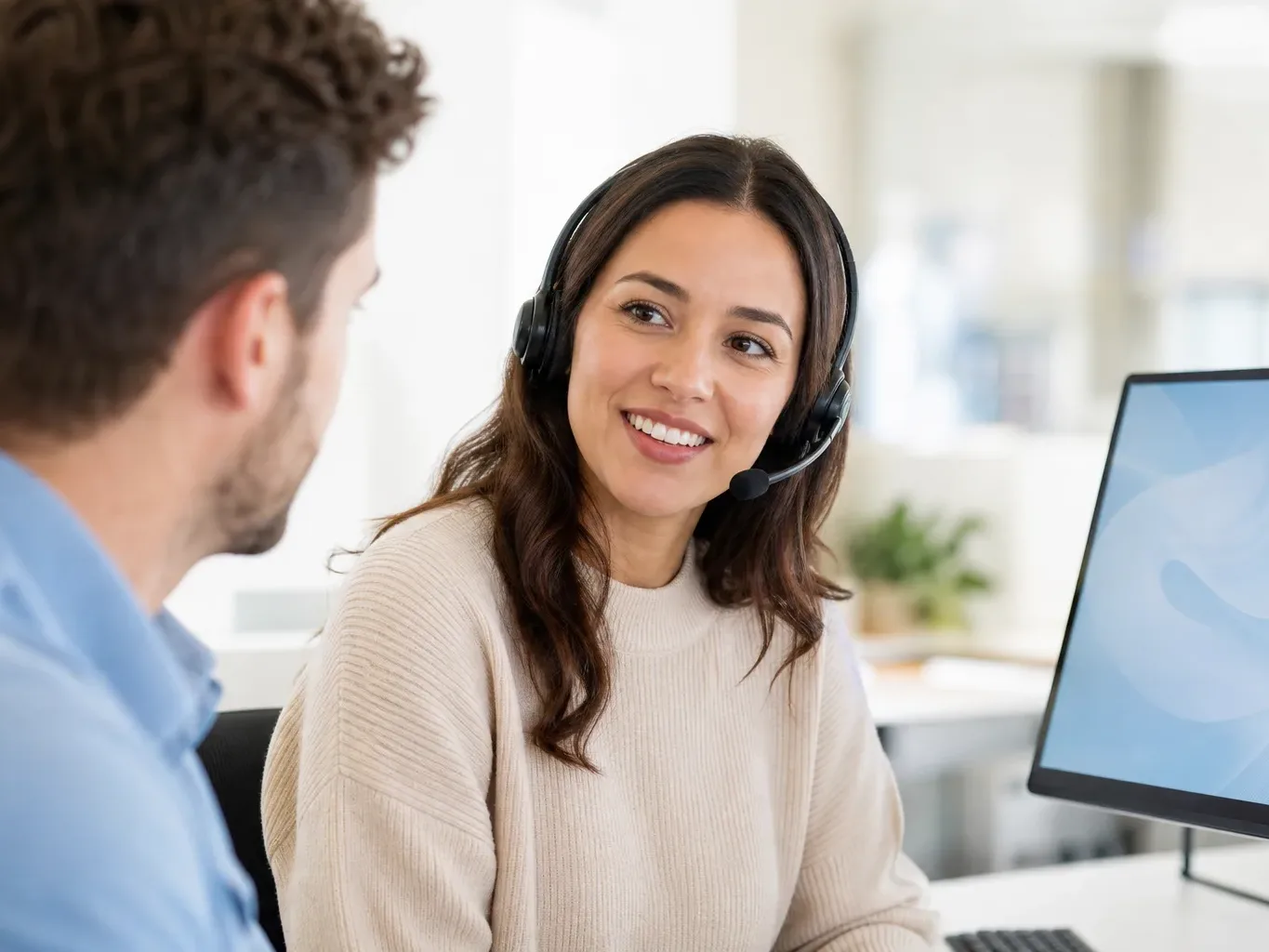 A customer service worker wearing a headset smiles while speaking with a colleague at an office desk beside a monitor