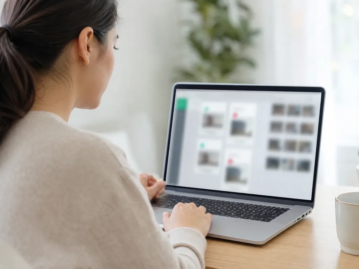 A person sitting at a desk uses a laptop displaying a blurred interface, with a small potted plant and a mug beside them in a bright home workspace