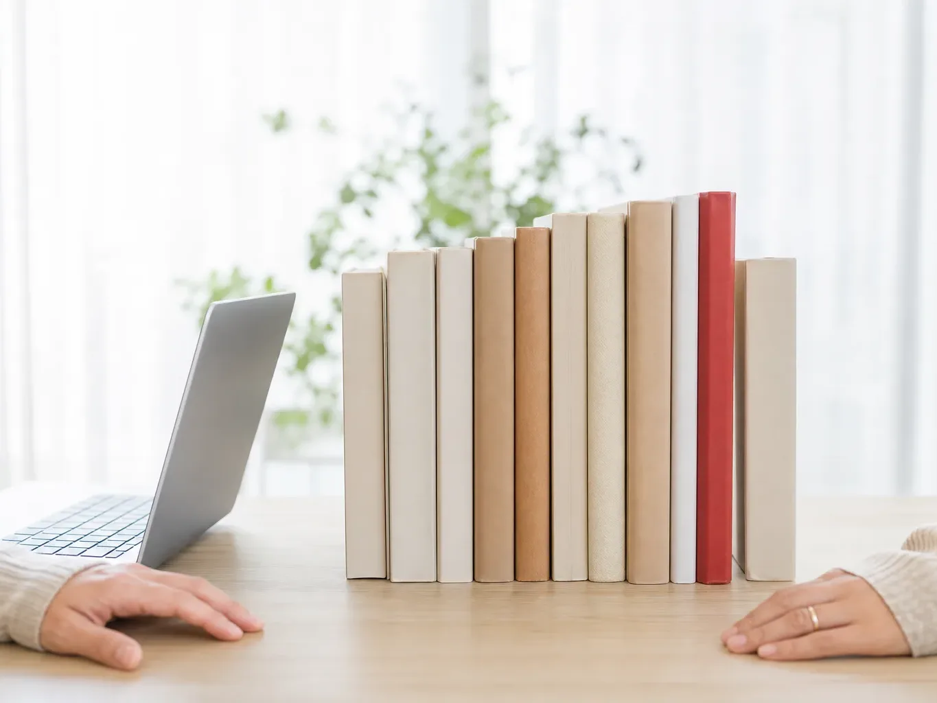 A person sits at a wooden desk with a laptop open to one side and a row of upright books between them and another person's hands resting on the opposite edge