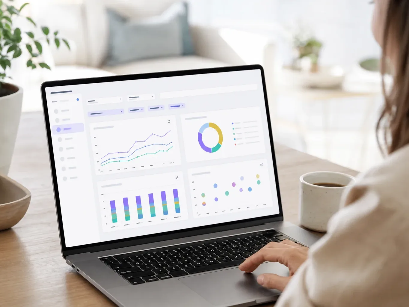 A person uses the trackpad of a laptop displaying a data analytics dashboard with line charts, a donut chart, a bar chart, and a bubble chart, beside a coffee cup on a wooden desk